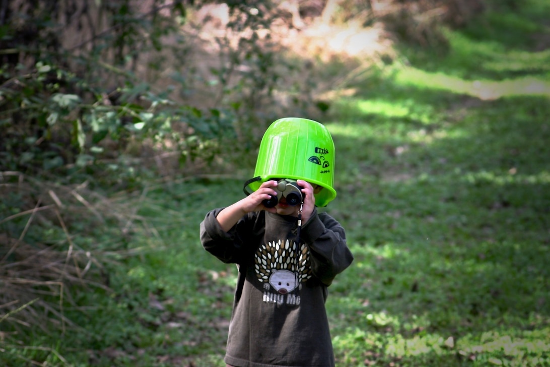 Kind met een felgroene bouwhelm en donkere kleding, die door een verrekijker kijkt in een natuurlijke buitenomgeving.
