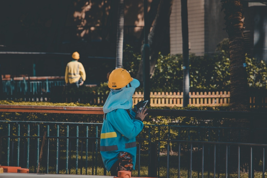 Kind in een felblauwe, goed zichtbare jas met reflecterende strepen en een oranje helm, staand bij een hek in een industriële omgeving.